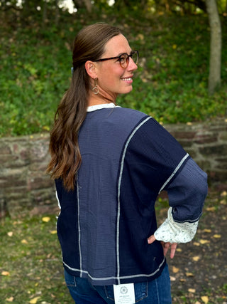 A woman wearing a navy blue textured long-sleeve top with white contrast stitching and white lace cuffs, photographed outdoors in a garden setting.