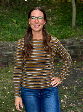 A woman with long brown hair and glasses stands outdoors wearing a long-sleeve crewneck shirt with horizontal brown, olive, and white stripes, paired with blue jeans.