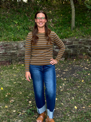A woman wearing a brown and tan striped long-sleeve knit top, blue jeans with rolled cuffs, and brown leather sandals, standing outdoors in front of a stone wall and green foliage.