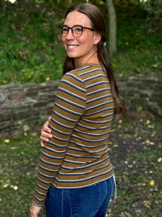 A woman wearing a brown, navy, and white striped long-sleeve knit top and blue jeans, photographed outdoors in a park setting.