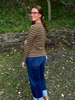 A woman wearing a brown and beige striped long-sleeve top and blue jeans stands outdoors in a wooded area, looking back over her shoulder.