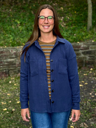 A woman with glasses and long brown hair stands outdoors wearing a navy blue button-up overshirt over a brown and beige striped shirt, paired with blue jeans. The background shows a blurred green garden setting.