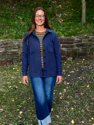 A woman wearing a navy blue button-up overshirt over a brown and yellow striped shirt, paired with blue jeans, standing outdoors in a grassy area with a stone wall behind her.