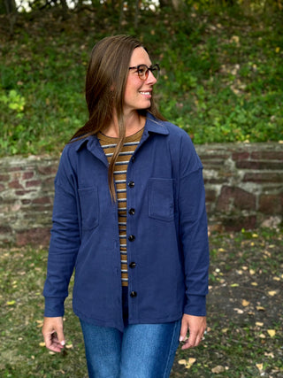 A woman wearing a navy blue button-up overshirt over a brown and white striped shirt, paired with blue jeans, standing outdoors in front of a stone wall and green foliage.