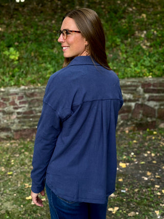 A woman with long brown hair and glasses, wearing a navy blue long-sleeve button-up shirt and dark blue jeans, standing outdoors in front of a stone wall and green foliage.