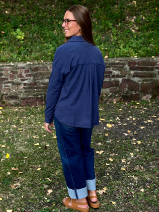 A woman wearing a navy blue button-up shirt, dark blue jeans with light blue cuffs, and brown sandals, standing outdoors in front of a brick wall and green foliage.