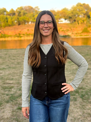 A woman with long brown hair and glasses stands outdoors in front of a lake and trees, wearing a black button-front vest over a black-and-white striped long-sleeve shirt and blue jeans.