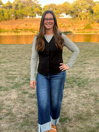 A woman with long brown hair and glasses stands outdoors in front of a lake and trees, wearing a black button-front vest over a black-and-white striped long-sleeve shirt and blue jeans.