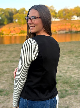 A woman wearing a black vest over a black and white striped long-sleeve shirt, standing outdoors in a grassy park with trees and water in the background.
