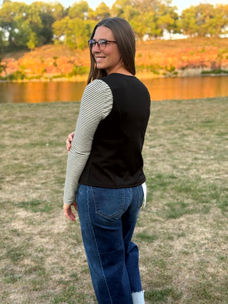 A woman wearing a black vest over a black and white striped long-sleeve shirt, blue jeans, and white socks, standing outdoors in a grassy field with a lake and trees in the background.