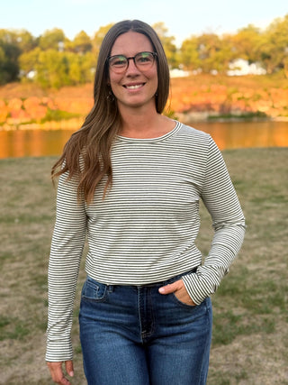 A woman with long brown hair and glasses stands outdoors in front of a lake and trees, wearing a long-sleeve crewneck top with thin black and white horizontal stripes and blue jeans.