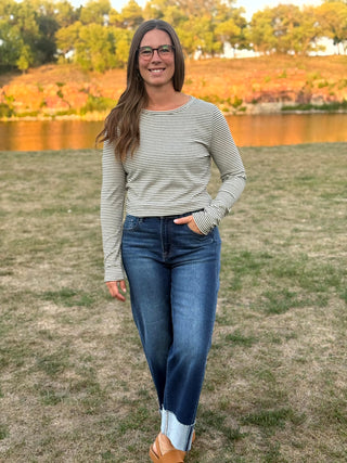 A woman wearing a light gray and white striped long-sleeve top, dark blue jeans, and tan loafers standing on a grassy lawn with a lake and trees in the background.