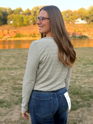 A woman with long brown hair and glasses, seen from behind, wearing a beige and black horizontally striped long-sleeve shirt and blue jeans, standing outdoors in a grassy park with a lake and trees in the background.