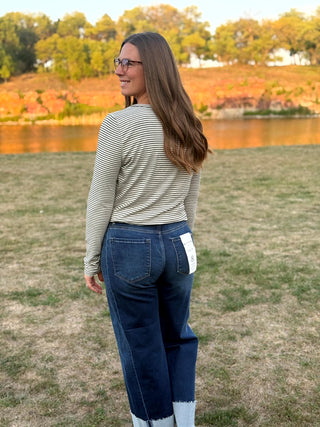 A woman wearing a beige and white striped long-sleeve top and dark blue jeans stands outdoors on a grassy field with a lake and trees in the background.