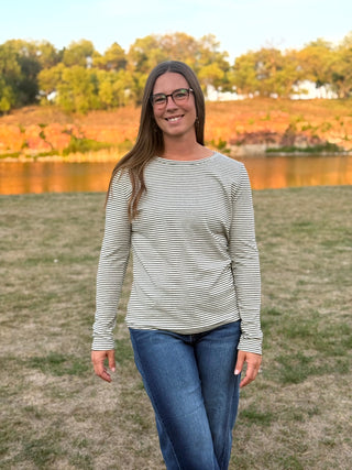 A woman with long brown hair and glasses stands outdoors in front of a lake and autumn trees, wearing a long-sleeve top with thin horizontal black stripes on a light beige or off-white base, paired with blue jeans.