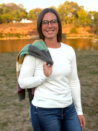 A woman wearing glasses, a white long-sleeve shirt, and blue jeans stands outdoors in front of a lake with trees in the background, holding a green, gray, and brown striped knit blanket over her shoulder.