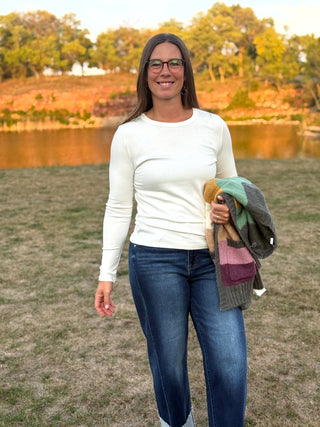 A woman wearing glasses, a white long-sleeve top, and blue jeans stands outdoors in a grassy field with a lake and trees in the background, holding a folded knit sweater.