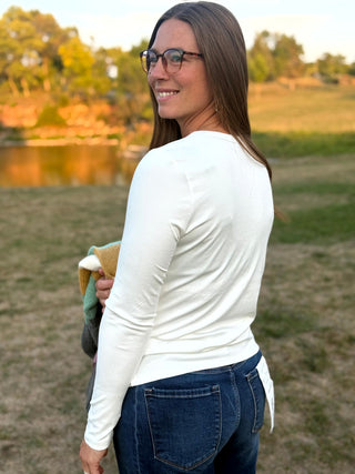 A woman wearing a fitted white long-sleeve top and blue jeans stands outdoors in a grassy field near a lake at sunset, holding a towel.