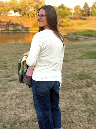 A woman wearing a white long-sleeve top and dark blue jeans stands outdoors in a grassy field near a lake, holding a folded multicolor knit blanket.