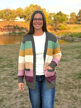 A woman wearing a colorful striped knit cardigan over a white t-shirt and blue jeans, standing outdoors in a grassy field with trees and a pond in the background.