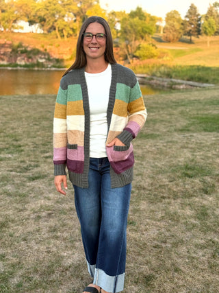 A woman wearing a multicolor striped knit cardigan over a white t-shirt and blue jeans, standing outdoors in a grassy field near a lake.
