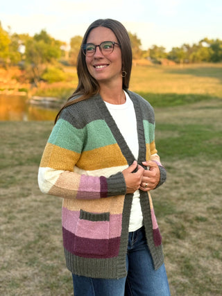 A woman wearing a chunky knit cardigan with horizontal stripes in olive green, mint green, mustard yellow, cream, dusty pink, and plum, standing outdoors in a grassy field.