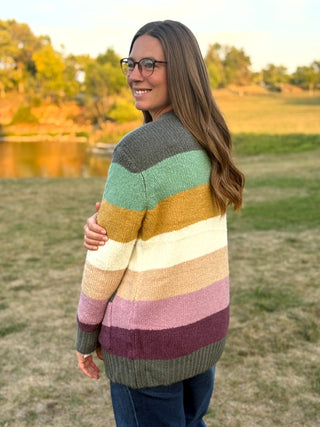 A woman wearing a fuzzy striped knit cardigan in muted earthy and pastel tones, standing outdoors in a grassy field near a lake at sunset.
