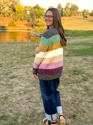 A woman wearing a colorful horizontally striped knit sweater, blue jeans, and sandals stands on a grassy field with trees and a pond in the background.