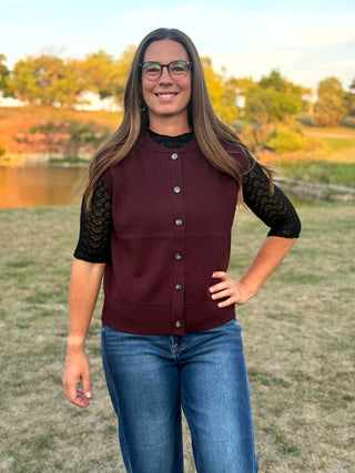 A woman wearing a sleeveless maroon button-up vest over a black lace-sleeve top and blue jeans, standing outdoors in a grassy field with trees and a pond in the background.