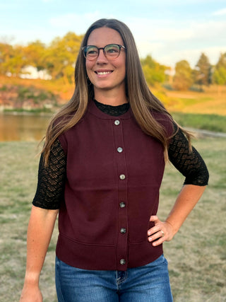 A woman wearing a sleeveless burgundy button-up vest over a black lace short-sleeve top and blue jeans, standing outdoors in front of a blurred park background.