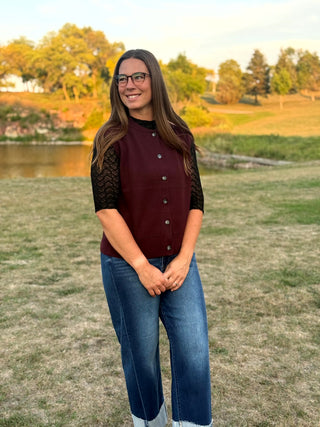 A woman wearing a maroon button-front cardigan with black lace sleeves and blue jeans, standing outdoors on a grassy field near a lake.