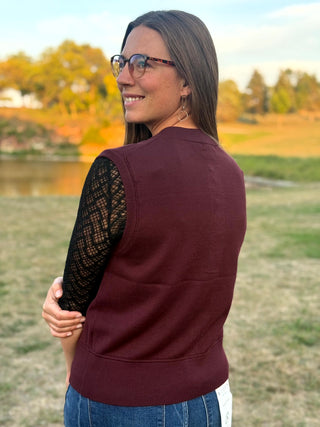 A woman wearing a sleeveless burgundy knit vest over a black lace long-sleeve top, photographed outdoors in a grassy park with trees and a pond in the background.