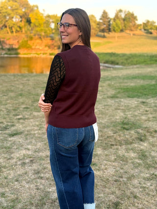 A woman wearing a sleeveless burgundy vest over a black lace-sleeve top and blue jeans, standing outdoors in a grassy field near a pond during golden hour.