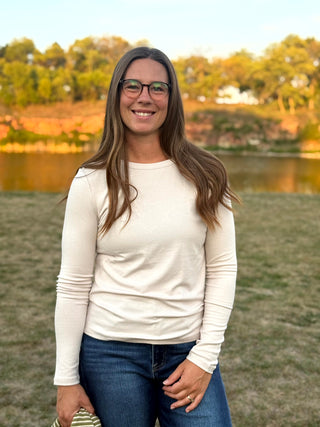 A woman with long brown hair and glasses stands outdoors in front of a lake and trees, wearing a fitted cream long-sleeve top and blue jeans.