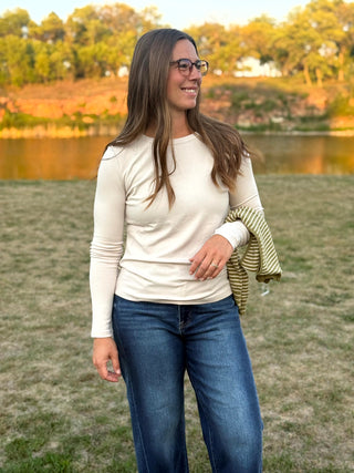 A woman wearing a cream long-sleeve crewneck top and blue jeans stands outdoors in a grassy field with a lake and trees in the background. She is holding a green and white striped knit sweater.