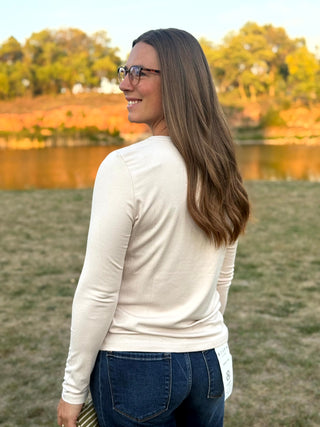 A woman wearing a cream long-sleeve crewneck top and blue jeans stands outdoors in a grassy field with a lake and trees in the background. She is holding a green and white striped knit sweater.