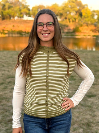 A woman with long brown hair and glasses stands outdoors in front of a lake and trees, wearing a sleeveless olive green and white striped button-up vest over a cream long-sleeve shirt and blue jeans.