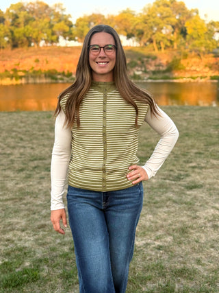 A woman wearing a sleeveless olive green and white striped zip-up vest over a cream long-sleeve shirt and blue jeans, standing outdoors in front of a grassy field and a lake with trees in the background.