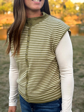 A woman wearing an olive green sleeveless button-front vest with thin white horizontal stripes over a cream long-sleeve shirt and blue jeans, photographed outdoors with a blurred natural background.