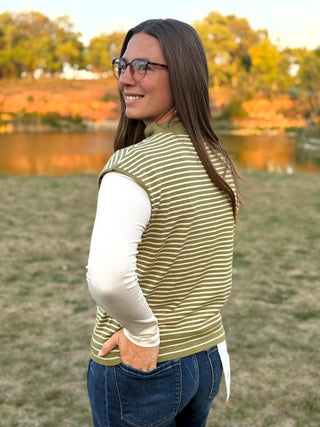 A woman wearing a sleeveless olive green and white striped knit vest over a white long-sleeve shirt, standing outdoors in a grassy field near a lake at sunset.