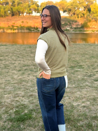 A woman wearing a sleeveless olive green and white striped knit vest over a white long-sleeve shirt, paired with dark blue jeans, standing outdoors on a grassy lawn near a lake.
