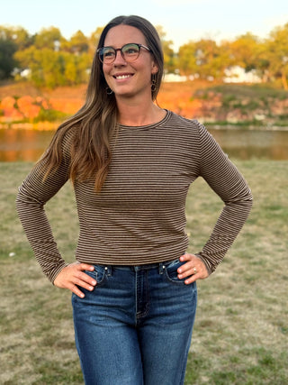 A woman wearing a brown and beige horizontally striped long-sleeve top and blue jeans, standing outdoors in a grassy park with trees and a lake in the background.