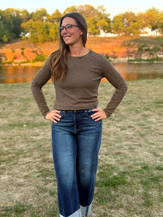 A woman wearing a brown and beige horizontally striped long-sleeve top and blue jeans stands outdoors on a grassy field with trees and water in the background.