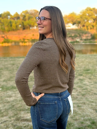 A woman with long brown hair and glasses, wearing a brown and beige horizontally striped long-sleeve top and blue jeans, standing outdoors in a grassy field with a lake and trees in the background.