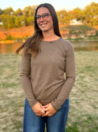 A woman with long brown hair and glasses stands outdoors wearing a brown and beige horizontally striped long-sleeve shirt and blue jeans, with a blurred background of grass, water, and autumn trees.