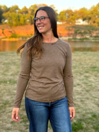 A woman wearing a brown and beige horizontally striped long-sleeve shirt and blue jeans, standing outdoors in front of a grassy field and a lake with autumn trees.