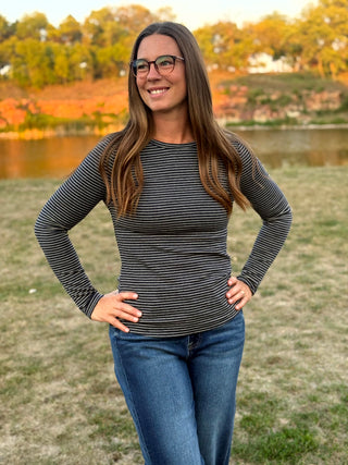 A woman with long brown hair and glasses stands outdoors on a grassy field with trees and water in the background, wearing a fitted long-sleeve top with thin dark horizontal stripes and blue jeans.
