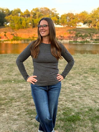 A woman with long brown hair and glasses stands outdoors in a grassy park near a lake, wearing a dark gray and black horizontally striped long-sleeve top and blue jeans.