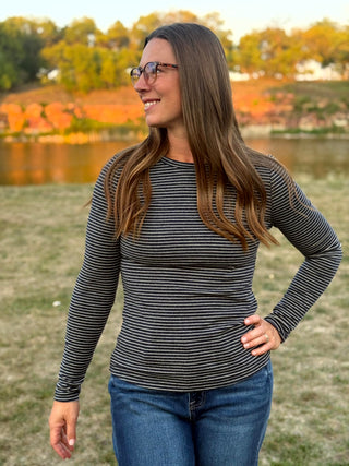 A woman wearing a black and gray striped long-sleeve shirt and blue jeans, standing outdoors in a grassy field with trees and a lake in the background.