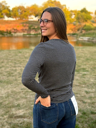 A woman wearing a black and gray striped long-sleeve top and blue jeans stands outdoors in a grassy field with a lake and autumn trees in the background.